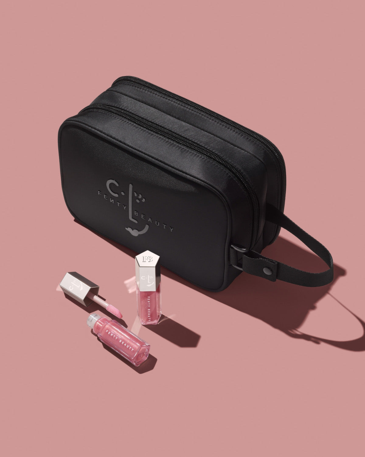 CLF makeup bag and two tubes of Gloss Bomb Universal Lip Luminizer in shade Candy Soda on a pink background.