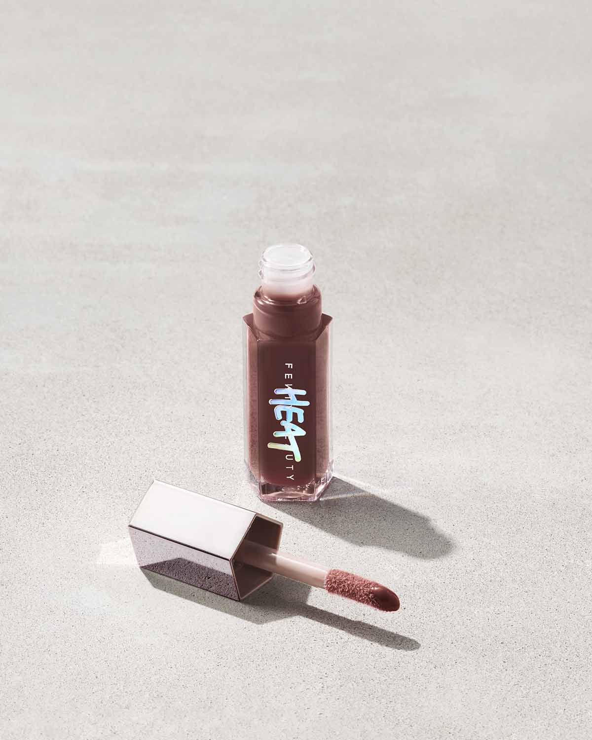 open tube of Fenty Beauty Gloss Bomb Heat Universal Lip Luminizer + Plumper in shade Hot Chocolit Heat with doe foot applicator on concrete background