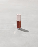 A close tube of Gloss Bomb Universal Lip Luminizer in the shade Hot Chocolit Fantasy on a grey background.