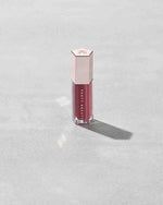 A closed tube of Gloss Bomb Universal Lip Luminizer in the shade RiRi on a grey background.