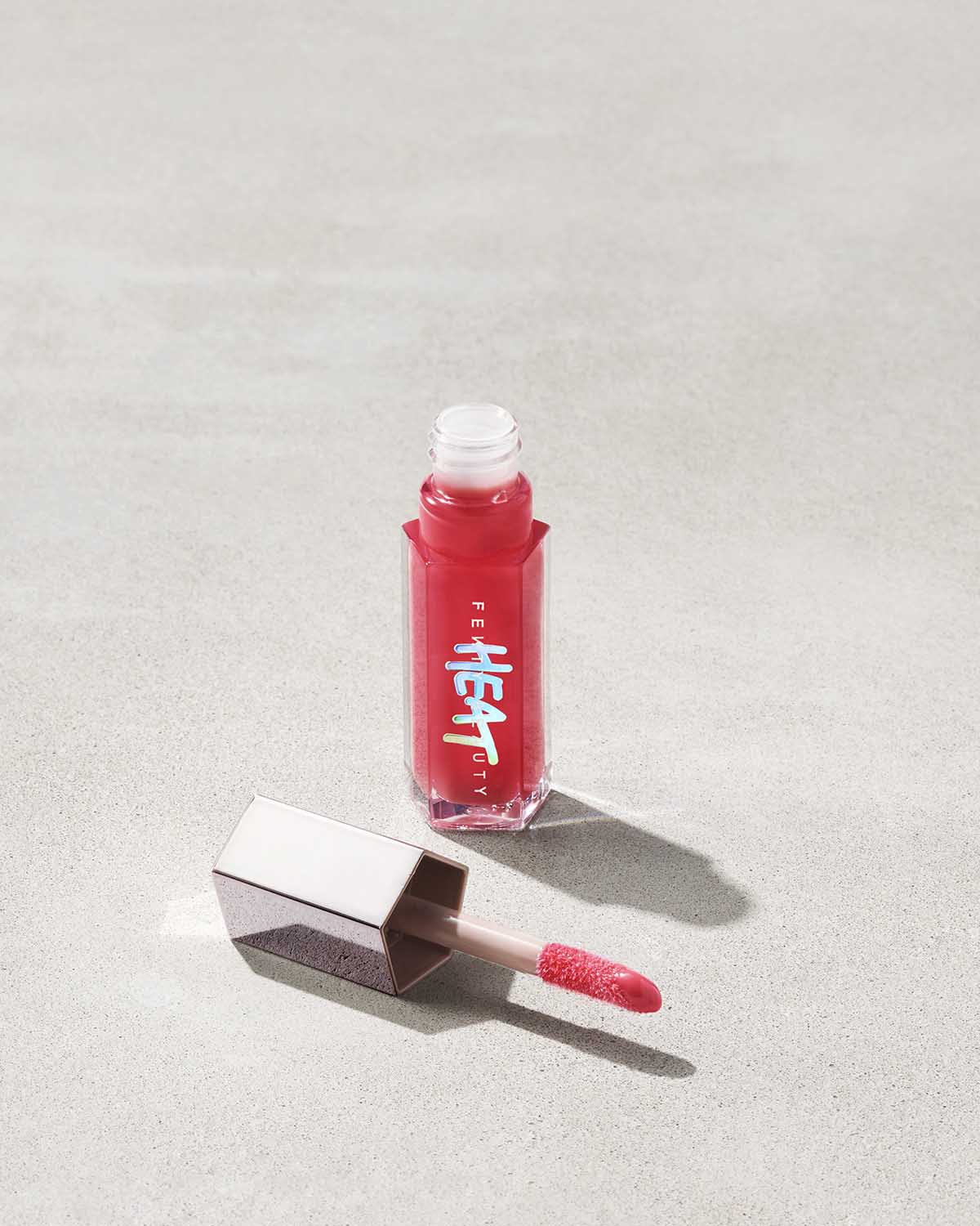 An opened tube of Gloss Bomb Heat lip gloss and plumpler in the shade Hot Cherry on a grey background.
