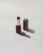 Open product shot of Gloss Bomb Stix Shimmer in shade Hot Chocolit on a concrete background.