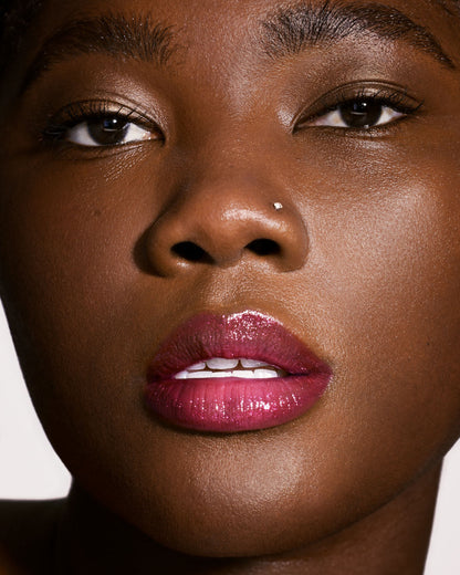 Close-up of a deep-toned model's face wearing Gloss Bomb Universal Lip Luminizer in the shade Fuchsia Flex.
