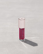 A closed tube of Gloss Bomb Universal Lip Luminizer in the shade Fuschia Flex on a grey background.