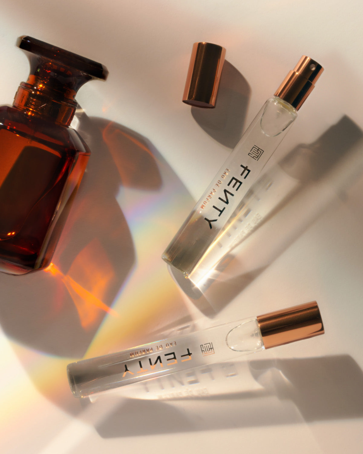 Two travel size perfume bottles of the Fenty Eau de Parfum next to the full size bottle, on a light background.